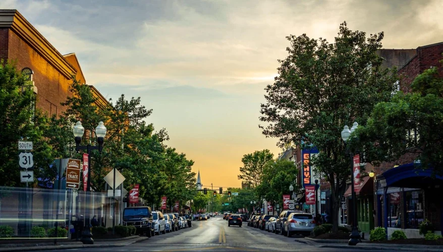 A street view of a charming downtown area, lined with trees and parked cars. The street leads towards the horizon with a warm sunset sky. Decorative banners are visible along the street, and buildings with various architectural styles are present on either side.