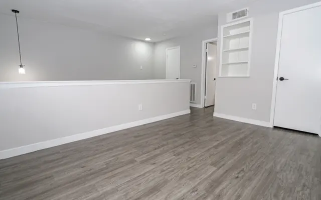 A modern interior space featuring light-colored walls, hardwood flooring, and minimalistic design elements. There is a pendant light hanging from the ceiling and an open area leading to a doorway on the right. The room is well-lit and spacious, giving a contemporary feel.