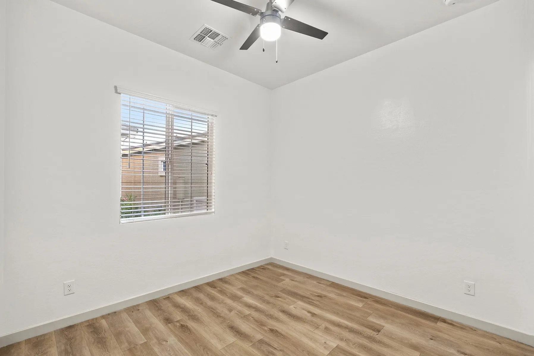 A minimalistic empty room with light-colored walls, a window with blinds, and wood flooring. A ceiling fan is visible.