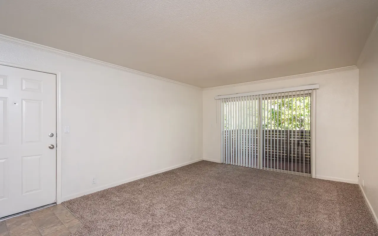 An empty living room with beige carpet and neutral-colored walls. There is a front door on the left and large sliding glass doors leading to a balcony on the right, adorned with vertical blinds.