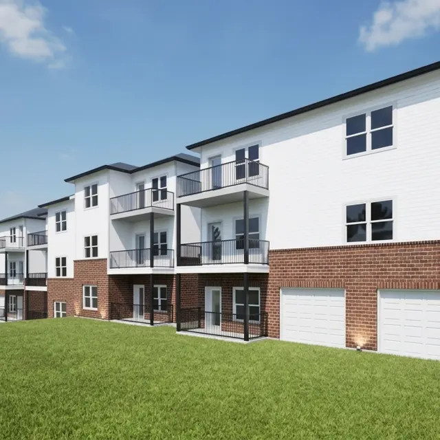 A modern apartment building featuring a combination of white siding and red brick. The structure has multiple levels, balconies, and garage doors, set against a green lawn and trees in the background under a clear blue sky.
