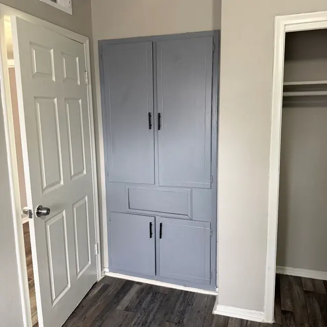 A room with a gray cabinet built into the corner, white walls, and a wooden floor. A door is visible beside the cabinet, leading to another area.