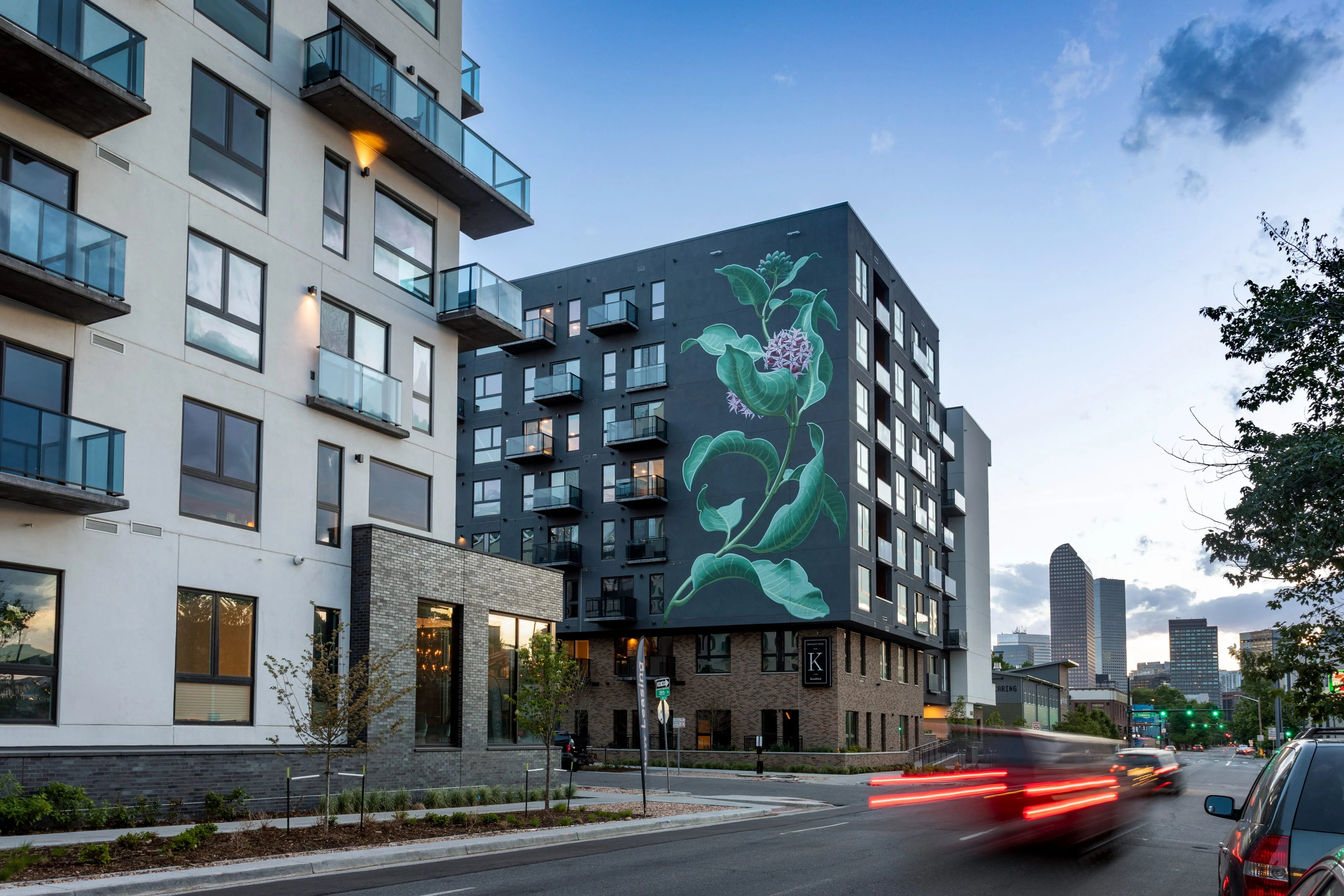 A modern urban residential building with balconies, featuring a large mural on one side. The mural displays a floral design. The scene is set in an urban street with car movement and city buildings in the background under a partly cloudy sky.