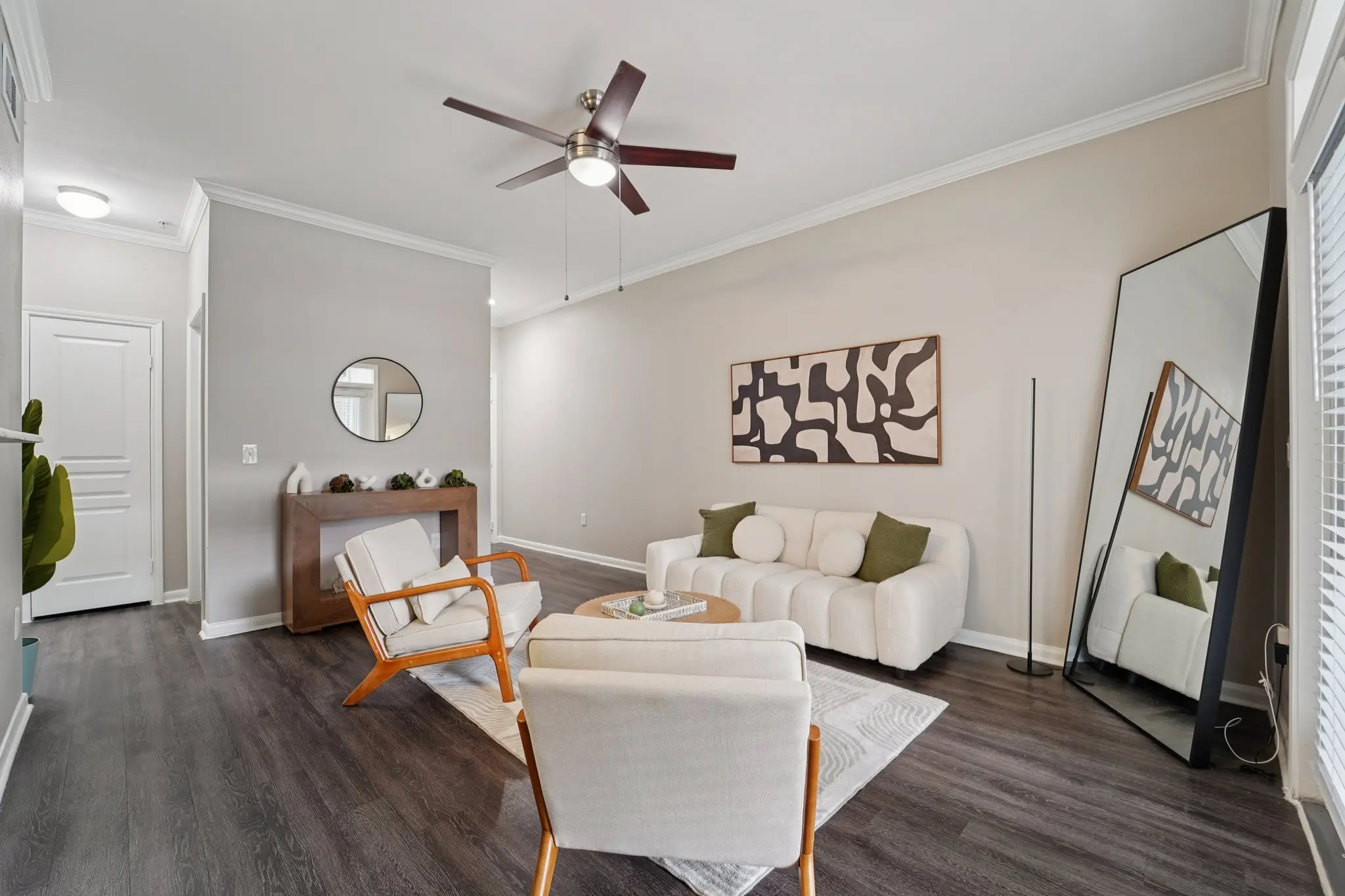 A modern living room with a light color scheme, featuring a white sectional sofa, two armchairs, a wooden console table, and a large mirror. A ceiling fan hangs above, and a piece of abstract art adorns the wall.