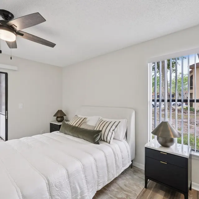 A modern bedroom featuring a comfortable bed with white bedding, a sleek nightstand with a lamp, a ceiling fan, and large windows allowing natural light, with a view of a patio.