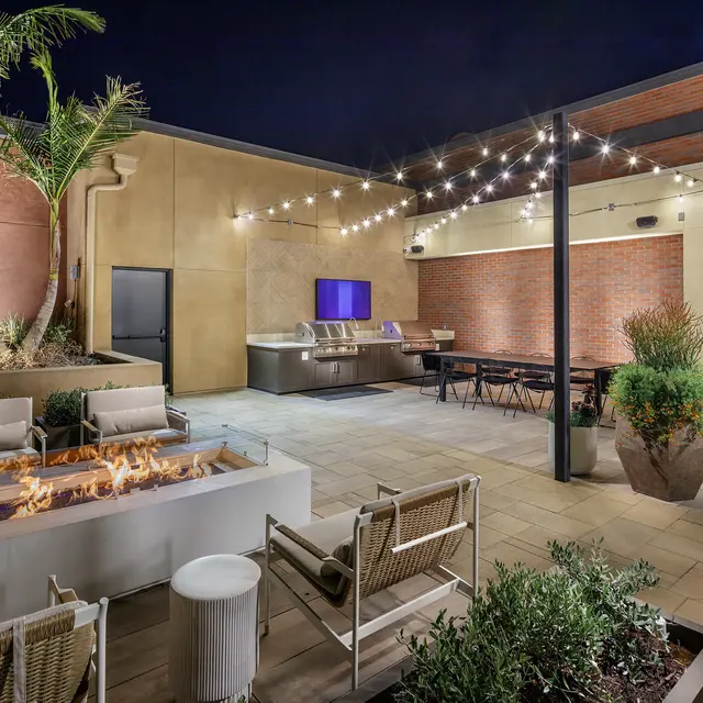 A modern rooftop lounge area at night featuring a fire pit, seating, string lights, and a grill.