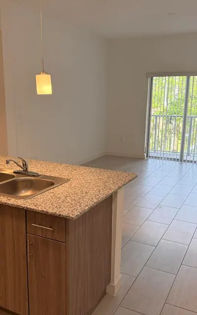 A modern apartment interior featuring a kitchen island with a sink, stainless steel appliances, and bright natural light coming from large glass doors leading to a balcony.