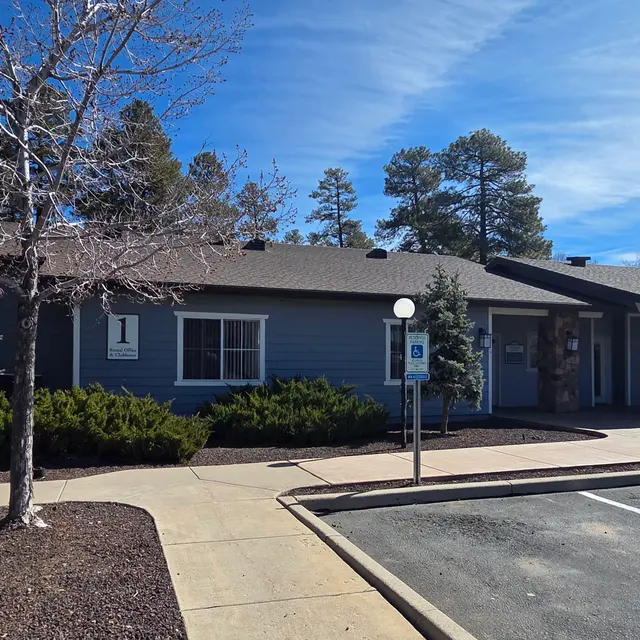 A blue building with a well-maintained landscape, featuring trees and a paved walkway leading to the entrance. The sky is clear with some clouds.