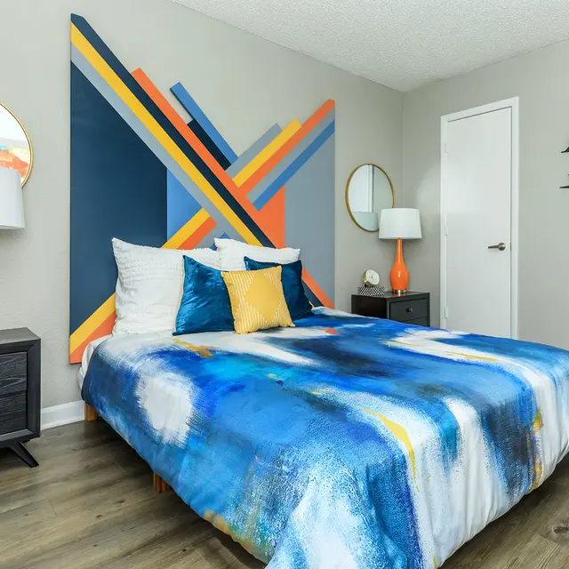 A modern bedroom featuring a blue, yellow, and white abstract patterned bedspread, decorative pillows, and a colorful geometric wall art. The room includes orange lamps, a round mirror, and a stylish black nightstand with decor items.