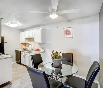 A modern kitchen and dining area featuring a glass table with black chairs, white cabinets, and a ceiling fan.