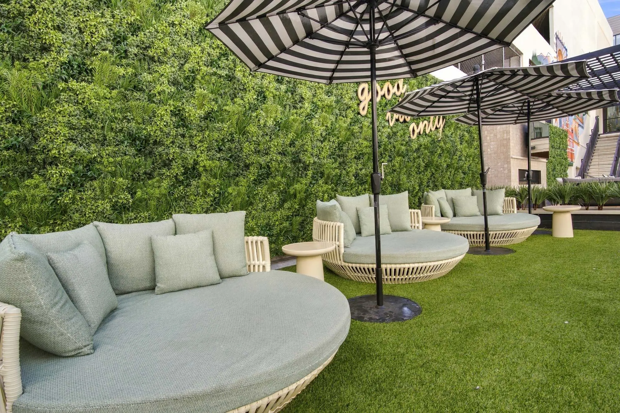 A cozy outdoor lounge area with round seating, decorative cushions, and striped umbrellas, surrounded by a green wall.