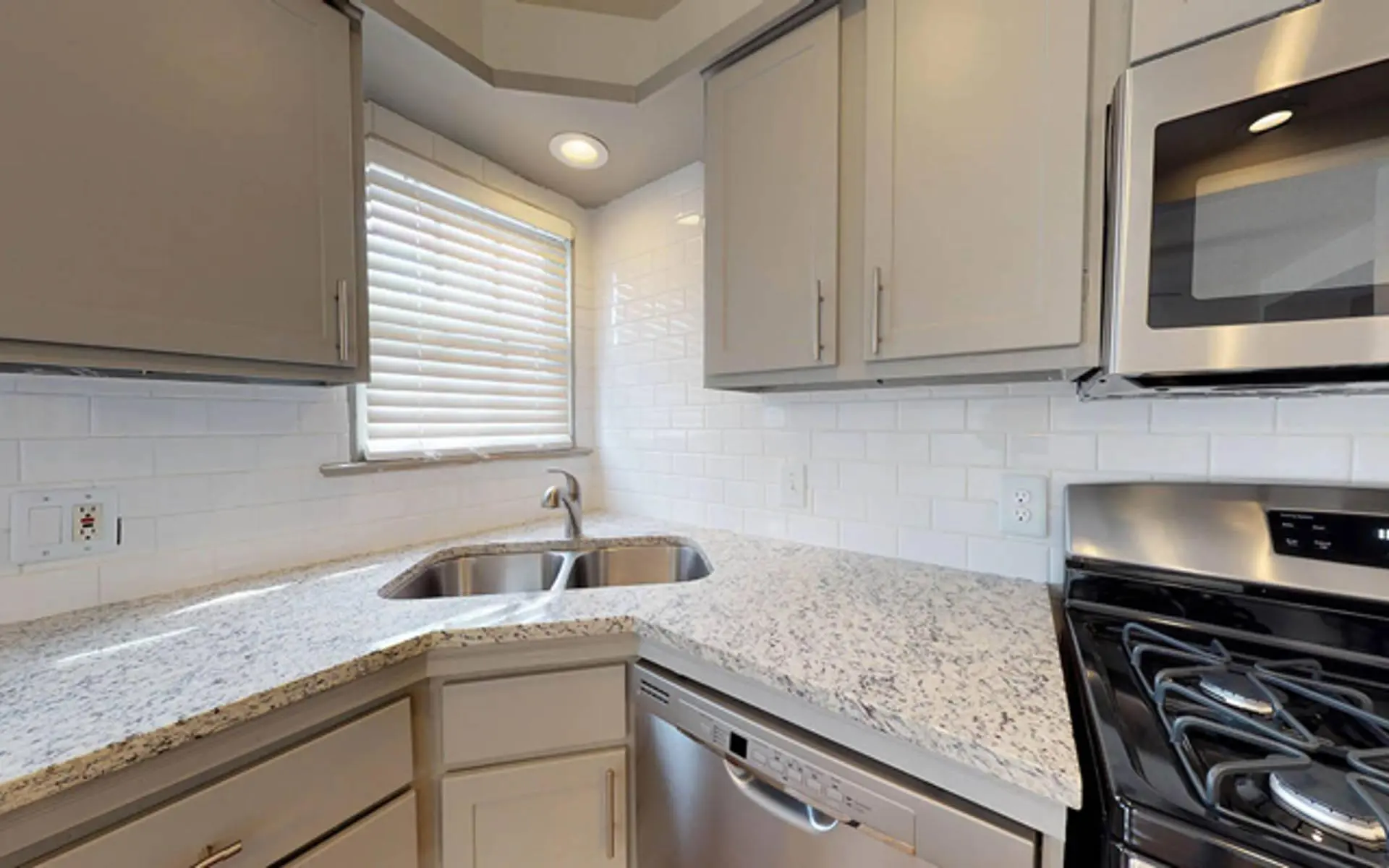 A modern kitchen featuring gray cabinetry, a white countertop with speckles, a double sink under a window with blinds, and a stainless steel stove and dishwasher.
