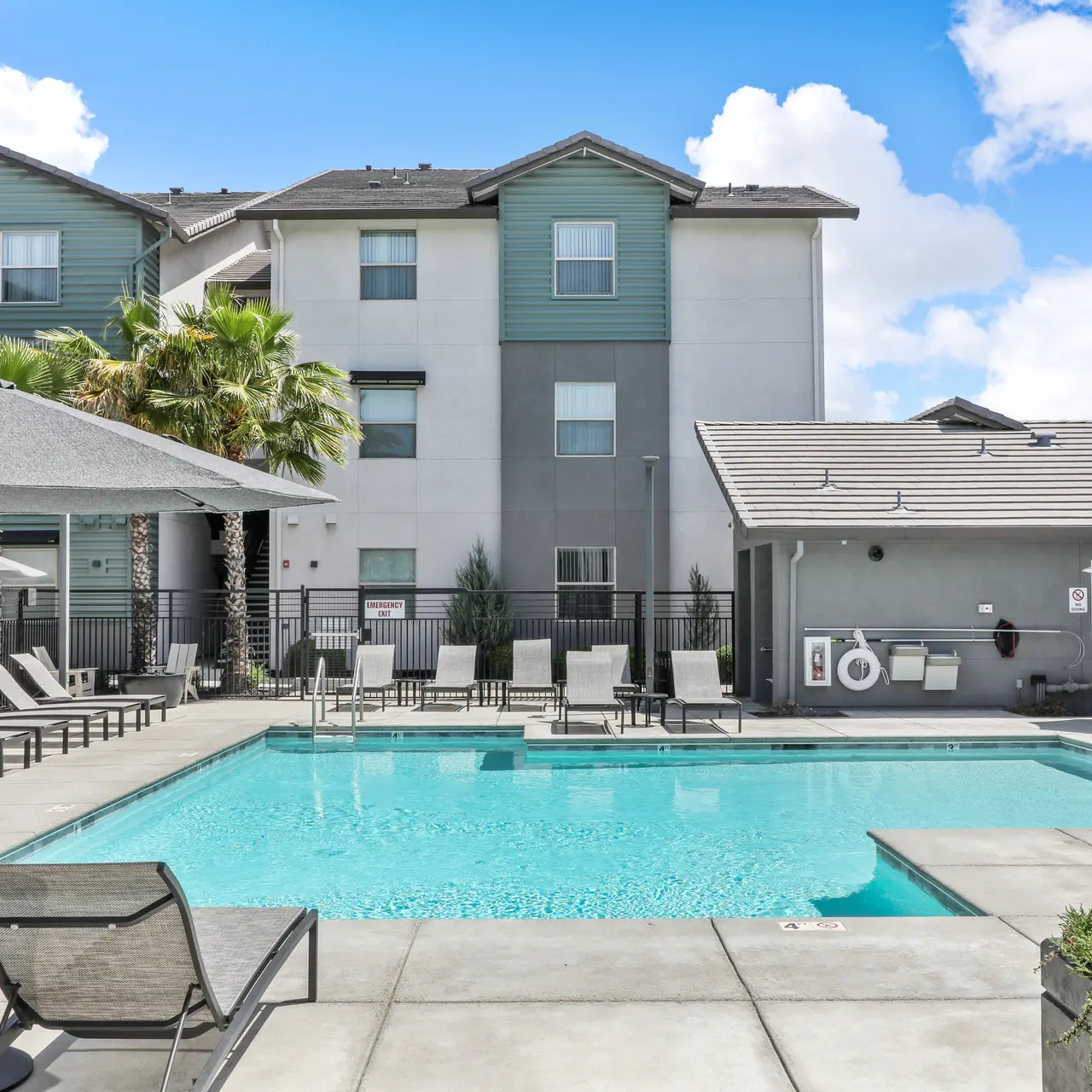 A swimming pool area at an apartment complex, featuring lounge chairs, umbrellas, and surrounding palm trees under a bright sky.