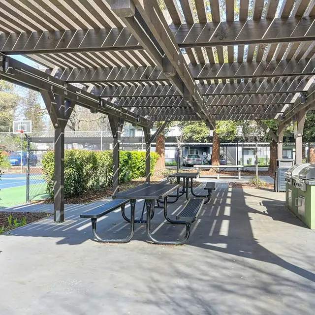 A shaded picnic area in a park with black picnic tables, a trash can, and a barbecue grill under a wooden pergola. In the background, there are tennis courts and lush green bushes.
