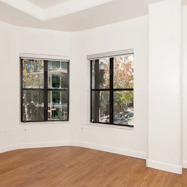 A modern, empty apartment corner with large windows showcasing city views and hardwood floors.