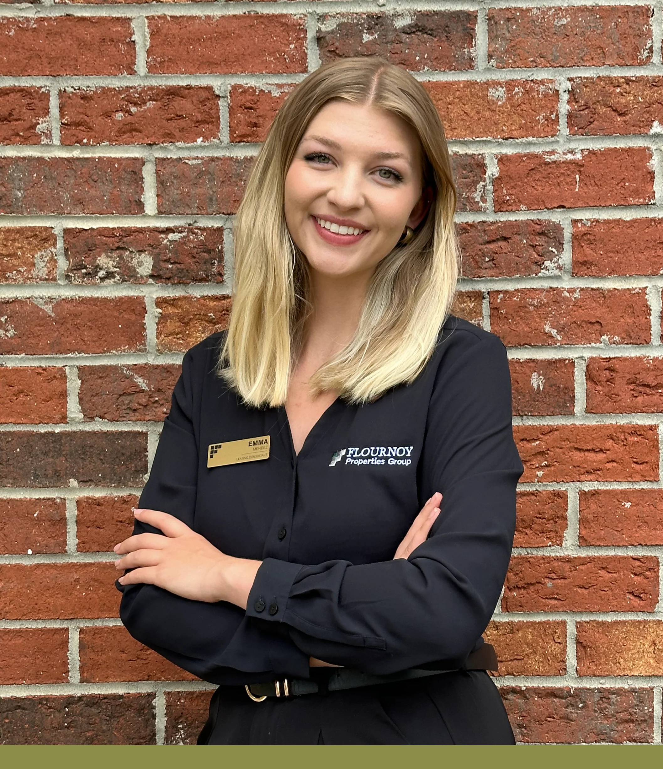 Real Estate Professional Photo A woman with long blonde hair wearing a navy blue shirt with a name tag and an emblem, standing with her arms crossed in front of a brick wall.