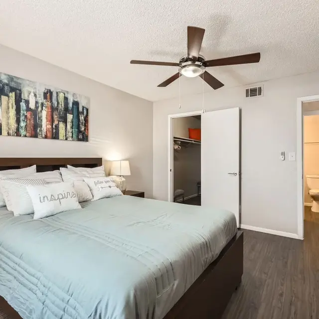 A modern bedroom featuring a large bed with light blue bedding, decorated with two bedside lamps and a wall art of a city skyline. A closet space is visible, and a bathroom is accessible through an open door in the background.