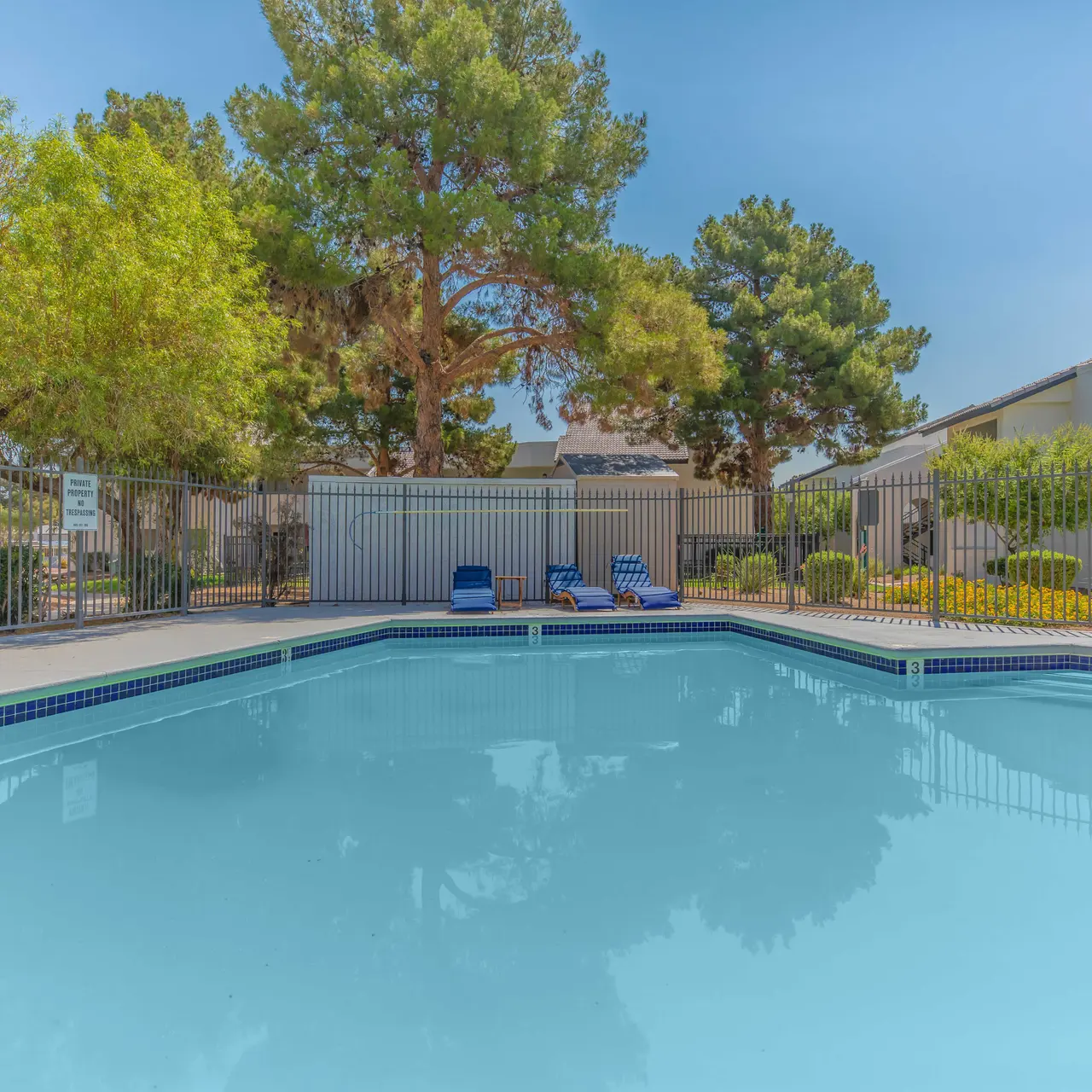 A serene swimming pool surrounded by greenery and residential buildings, with blue lounge chairs near the edge.