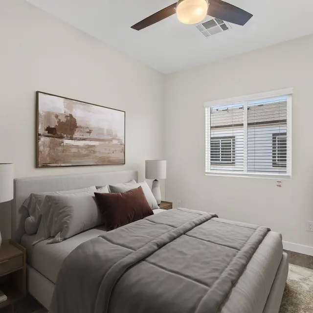 A modern bedroom featuring a queen-sized bed with gray bedding and two bedside tables with lamps. There's an abstract painting above the bed and a houseplant in the corner. A window provides natural light, and the room has light-colored walls and wood flooring.