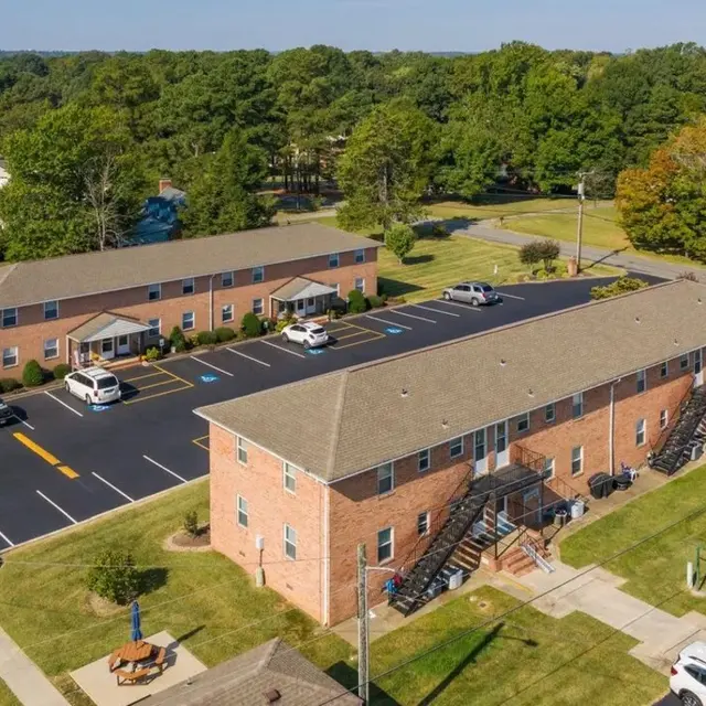 Aerial view of a small apartment complex consisting of two brick buildings with multiple units. One building has a series of stairs leading to its entrance. The parking lot is visible, with several parked cars, and there are grassy areas around the buildings.