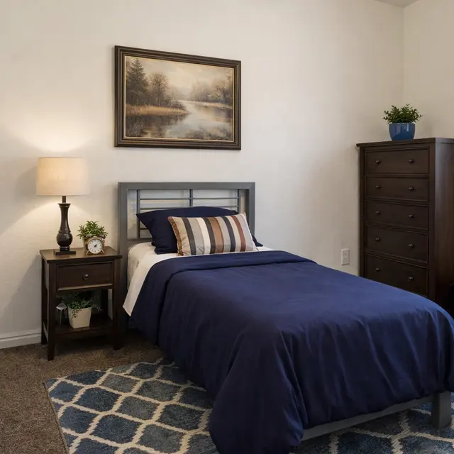 A cozy single bedroom featuring a bed with navy blue bedding, a bedside table with a lamp, and a dresser. There is an artwork on the wall and a plant on the table. A patterned rug covers the carpeted floor, and a woven basket is situated near the dresser.