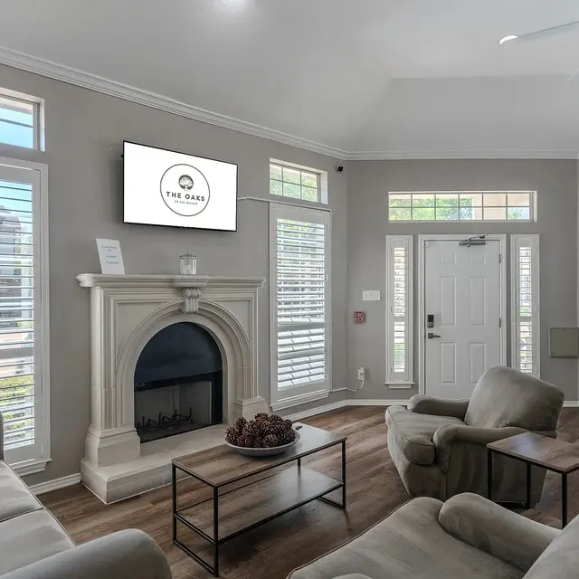 A modern living room with a fireplace, large windows, and light-colored walls. There are comfortable seating arrangements and a mounted television displaying a logo.