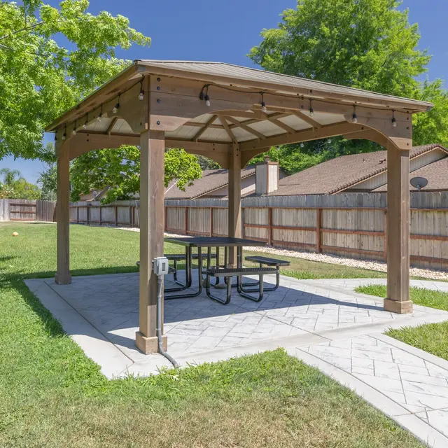 A wooden gazebo with a picnic table located in a grassy backyard area, surrounded by trees and residential buildings.
