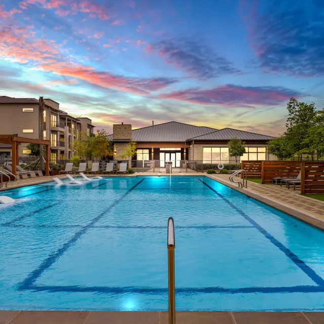 A luxurious swimming pool surrounded by lounge chairs, with modern apartment buildings in the background under a colorful sunset sky.