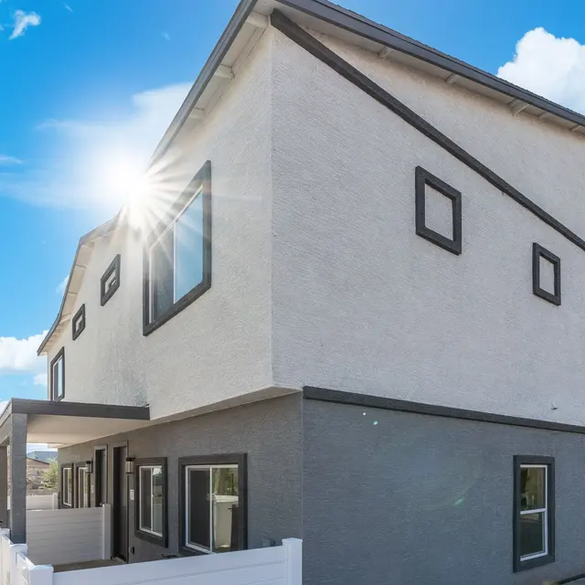 Contemporary Two-Story House A modern two-story house with a light-colored exterior featuring dark trim. The house is positioned at an angle with sunlight glimmering off one corner. The sky is blue with white clouds, and a white fence surrounds the property.