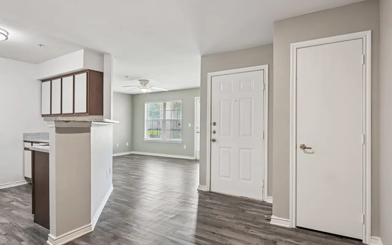 Interior view of a modern apartment showcasing a living area with hardwood flooring, a ceiling fan, and a front door entrance. A small kitchen area is visible to the left.