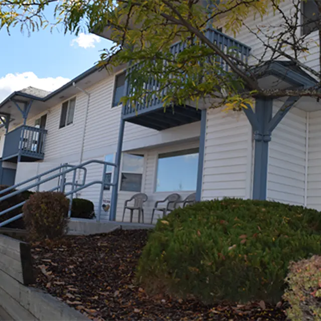 Exterior of a building featuring a light-colored façade, blue accents, and a landscaped front area with shrubs and seating.