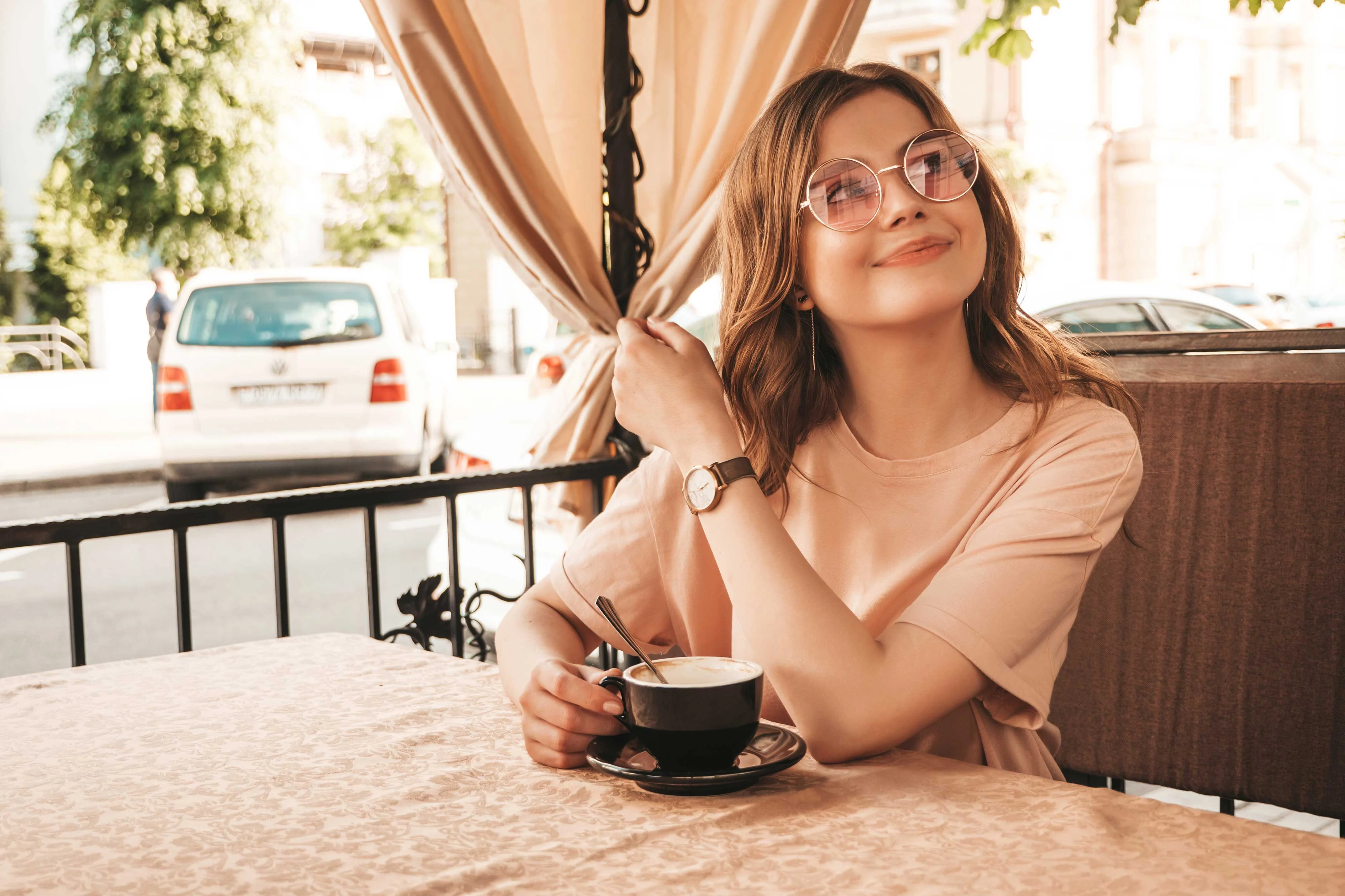 A woman with glasses holding a cup of coffee while sitting at a table in a cafe, with a light curtain draped nearby and a car visible in the background.
