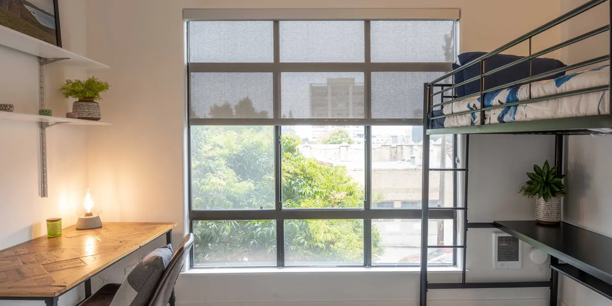 A cozy, modern study room featuring a bunk bed, a desk with a chair, and large windows allowing natural light to filter in. Green plants are placed on the desk and on the shelf, with an urban outdoor view visible through the window.