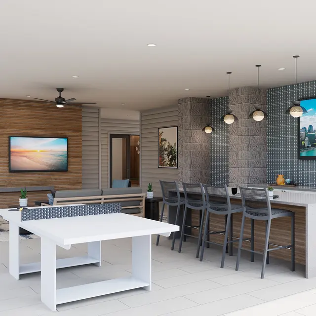 A modern outdoor kitchen area featuring a bar with high chairs, large screens displaying city views, and a dining table. The space has sliding doors leading to a corridor, light-colored flooring, and contemporary lighting fixtures.