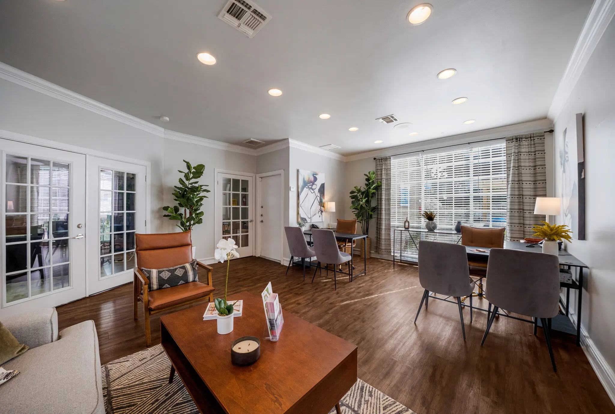A cozy living area featuring modern furniture, hardwood flooring, and large windows with natural light. There are two plants, a brown sofa, a wooden coffee table, and several chairs.