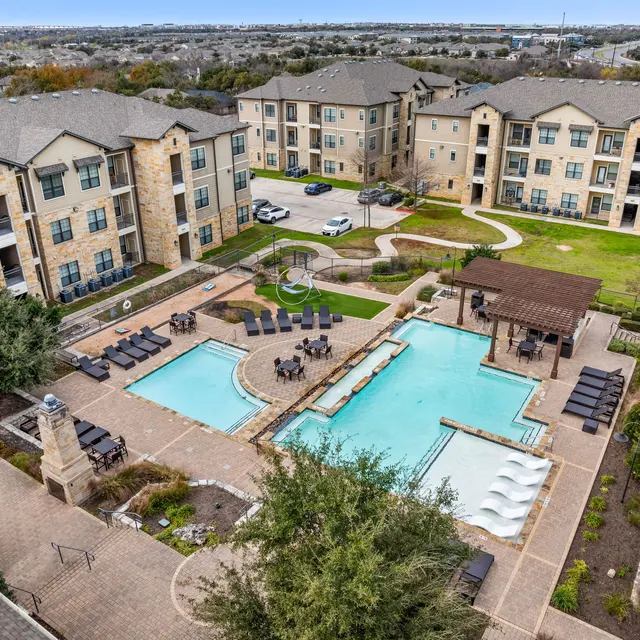 Aerial view of an apartment complex featuring a pool area with loungers, landscaped surroundings, and nearby buildings.