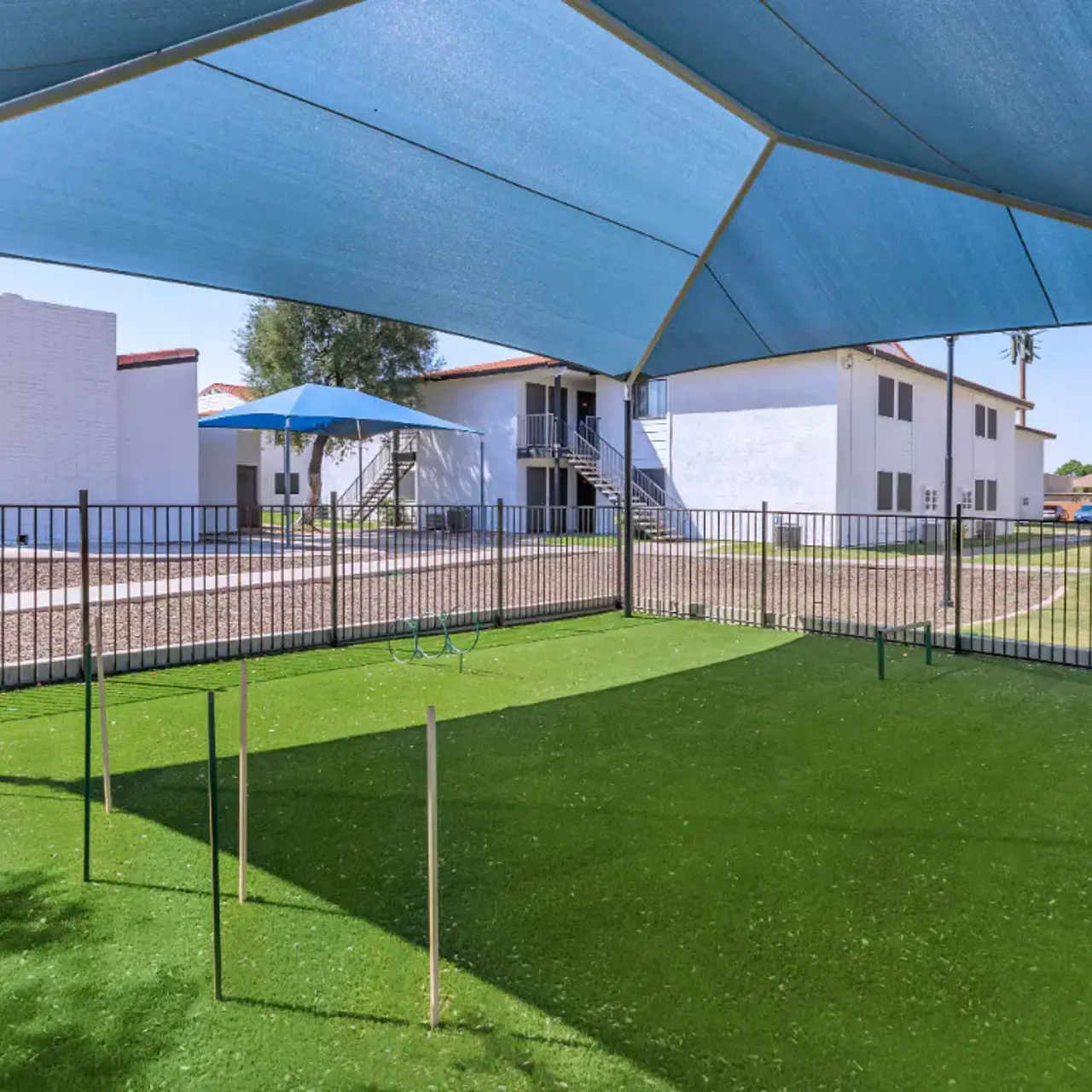 A shaded area with green artificial grass surrounded by a black fence, featuring white buildings in the background and a blue shade sail overhead.