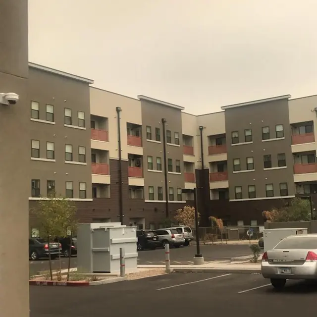 Modern Apartment Complex A modern apartment complex featuring multiple stories, parking spots, and several cars parked outside. The building has a mix of gray and beige colors, with some balconies visible. The sky appears overcast, providing a muted atmosphere.