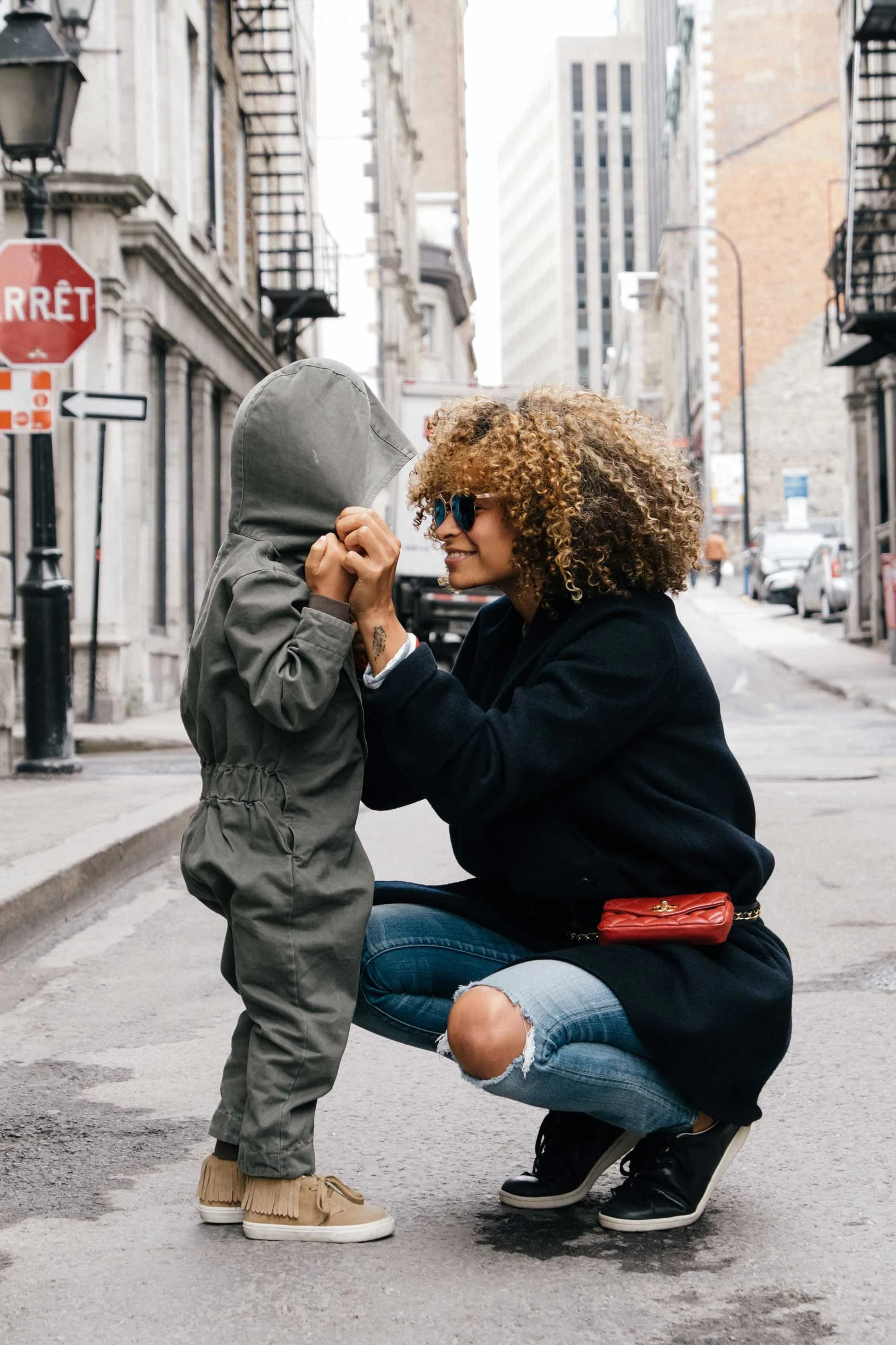 City Connection A woman with curly hair kneels down to interact with a young child wearing a green jumpsuit. They are in a city street with buildings in the background.