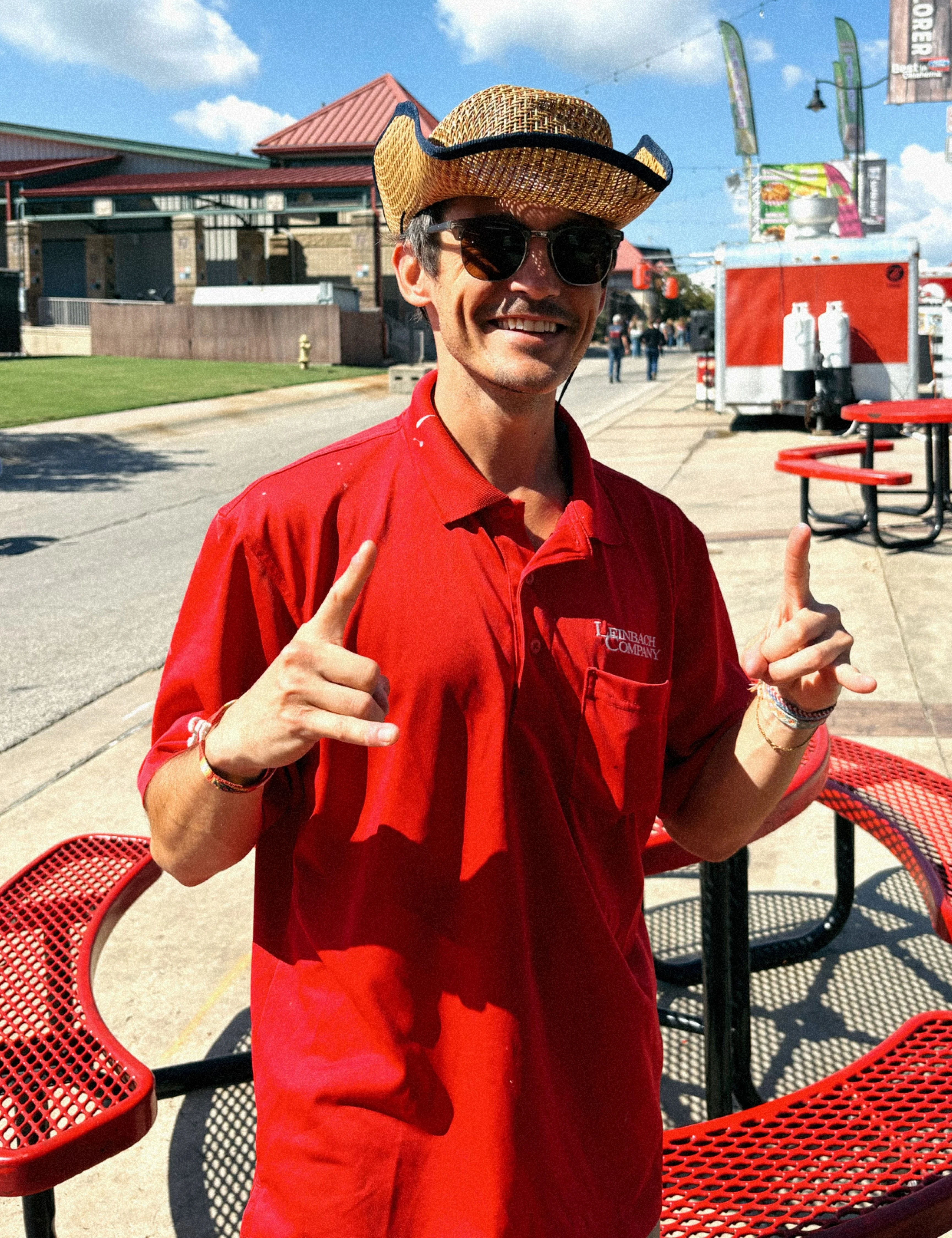 Man in Red Shirt at Food Festival A man wearing a red polo shirt and a straw cowboy hat stands outdoors, smiling and gesturing with his hands. There are tables and food trucks in the background.