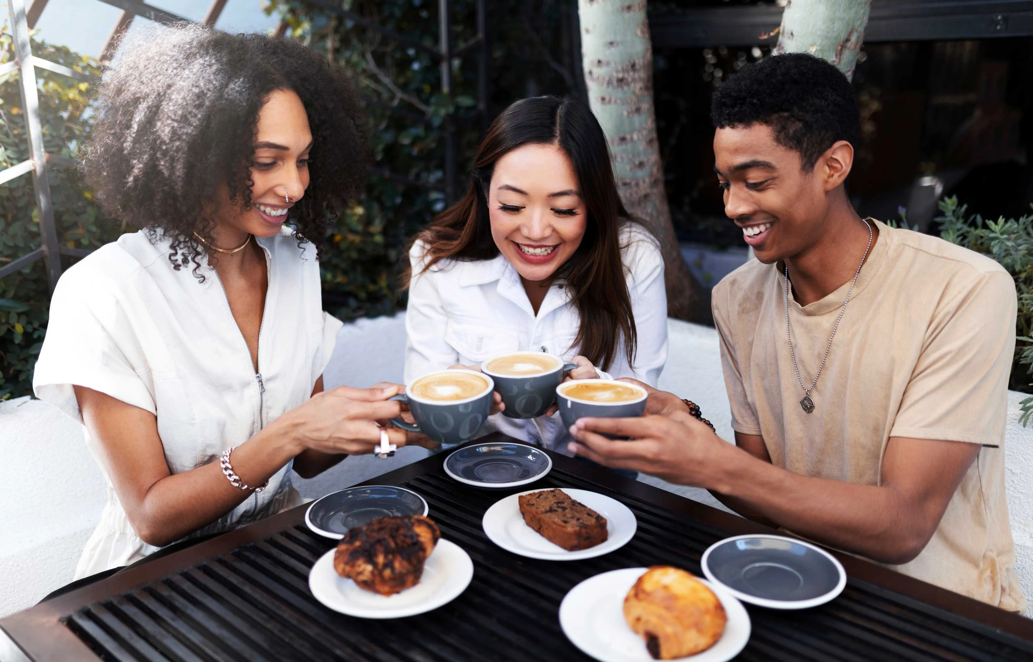 Three friends sitting at a table enjoying coffee and pastries, smiling and toasting their cups.