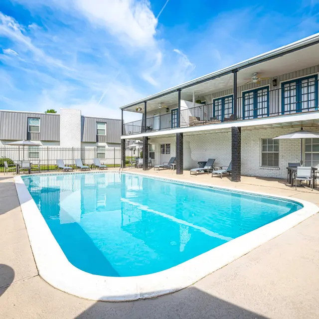 A well-lit swimming pool area featuring a large blue pool surrounded by lounge chairs and a two-story building with balconies overlooking the pool.