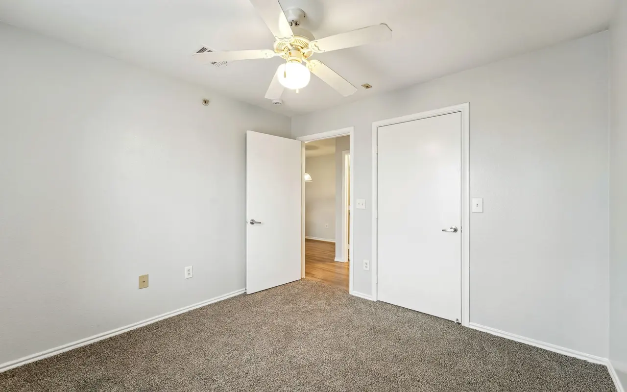 A light and airy empty room with beige carpet and white walls. A ceiling fan is visible. Two doors lead to adjoining spaces.