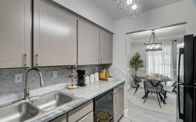 A modern kitchen featuring light-colored cabinets, granite countertops, and stainless steel appliances. In the background, there is a dining area with a round table and stylish chairs, illuminated by a suspended light fixture.