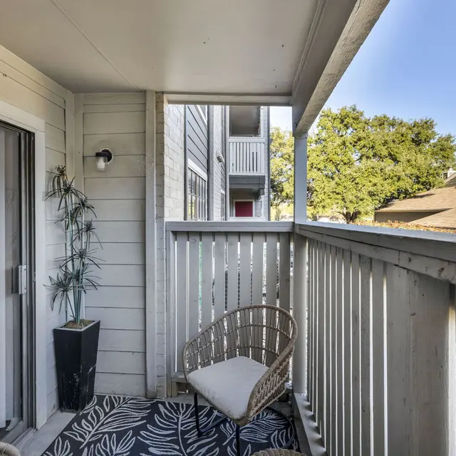 Cozy balcony with a rattan chair, potted plant, and patterned rug, overlooking nearby buildings and trees in daylight.