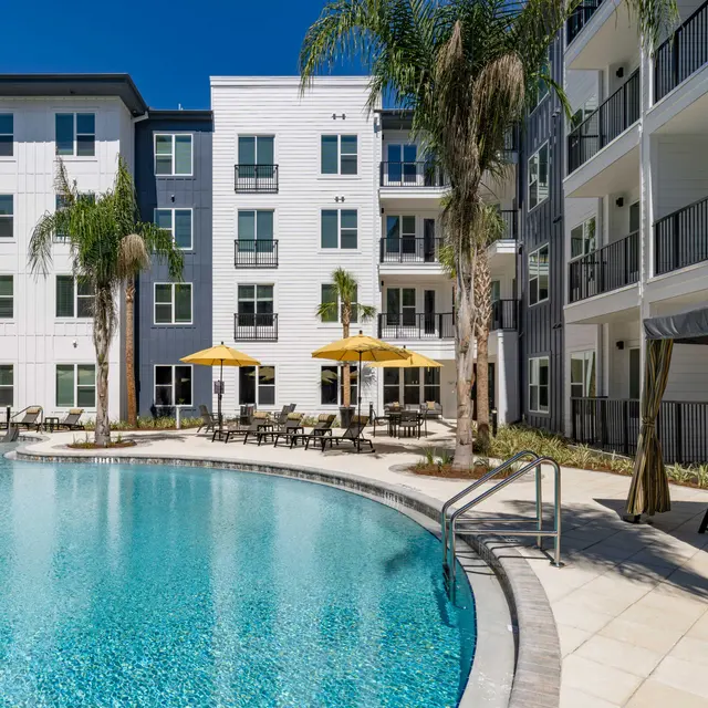 A luxurious apartment complex featuring a large swimming pool surrounded by lounge chairs and umbrellas, with modern buildings in the background.