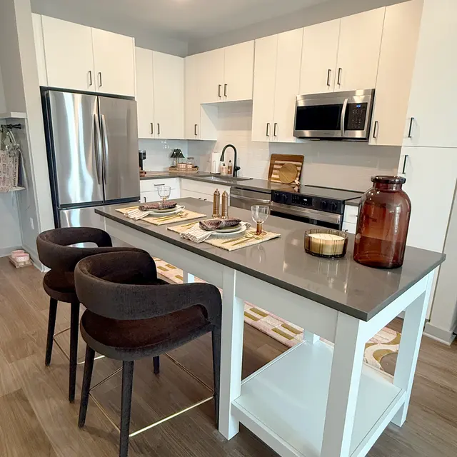 A modern apartment kitchen in Richmond VA featuring white cabinets, stainless steel appliances, and a gray countertop with four barstools on one side.