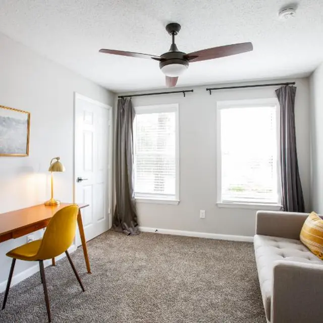 A cozy home office with a grey carpet, a wooden desk and a yellow chair, two windows with curtains, and a sofa with orange pillows.