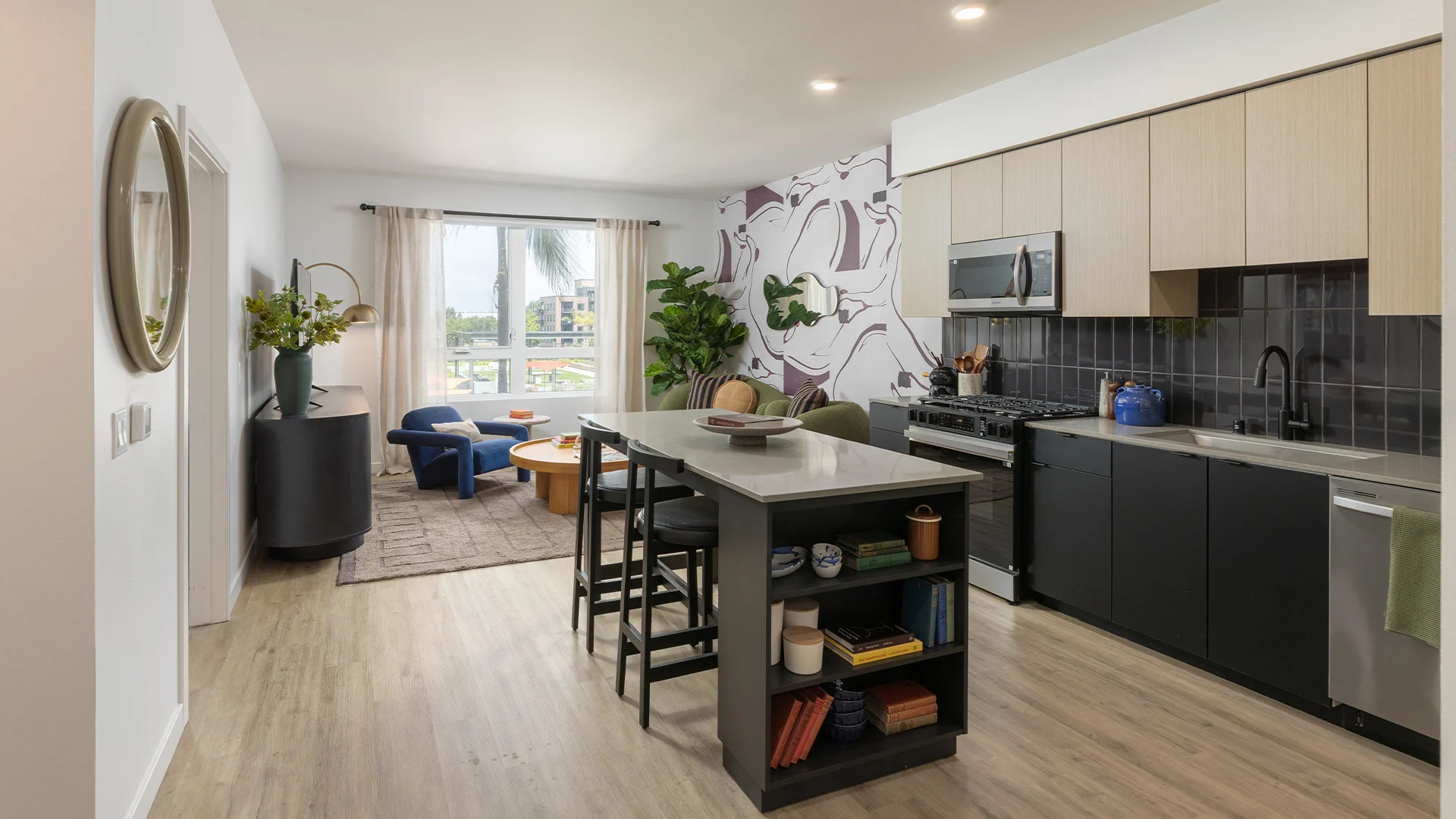 Modern Kitchen and Living Area A modern kitchen and living area featuring a spacious layout, light wood flooring, and contemporary decor. The kitchen includes sleek cabinetry and stainless steel appliances, while the living area has a colorful rug and a small seating arrangement near a large window.