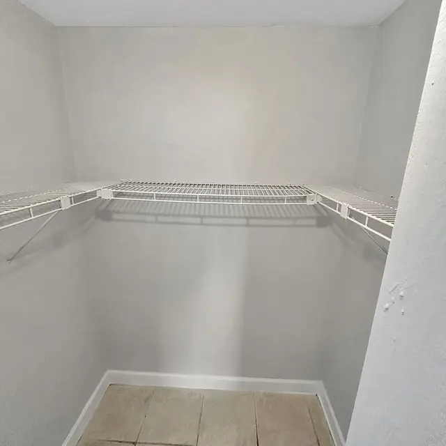 An empty closet featuring light gray walls and tiled flooring. There is a wire shelf mounted on one side with no items on it, creating a spacious but bare appearance.